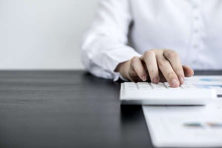 A woman uses a laptop and presses a white calculator, she is a company auditor, she checks documents from the laptop and calculates the numbers for accuracy. Financial audit conceptの写真素材