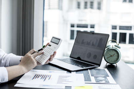A woman uses a laptop and presses a white calculator, she is a company auditor, she checks documents from the laptop and calculates the numbers for accuracy. Financial audit conceptの写真素材