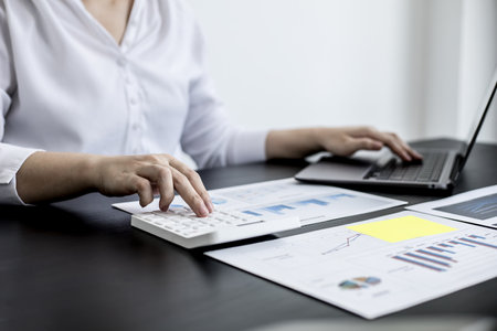 A woman uses a laptop and presses a white calculator, she is a company auditor, she checks documents from the laptop and calculates the numbers for accuracy. Financial audit conceptの写真素材