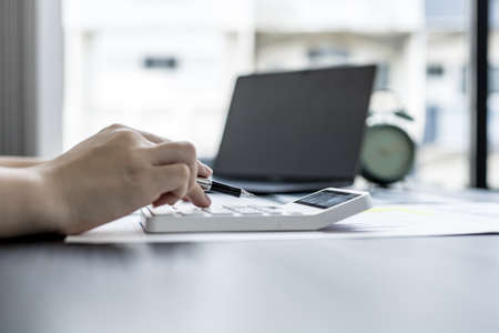 A woman uses a laptop and presses a white calculator, she is a company auditor, she checks documents from the laptop and calculates the numbers for accuracy. Financial audit conceptの写真素材