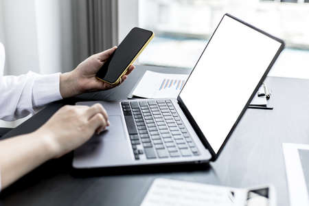 A woman holding a smartphone and using a laptop uses a smartphone. Chat with friends through instant messaging to discuss business together and use your laptop to find information on the web.の写真素材