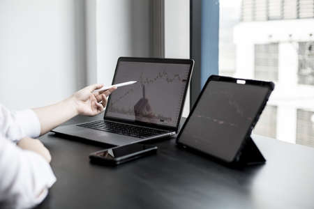 A stock investor holding a pen pointing to a laptop screen opens a stock chart program to read stock...