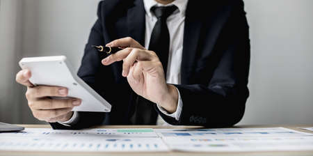 A man with a pen pointing to a white calculator and documents are placed on the table, a businessman in a black suit is sitting to check information from documents, he is a company executive.の写真素材