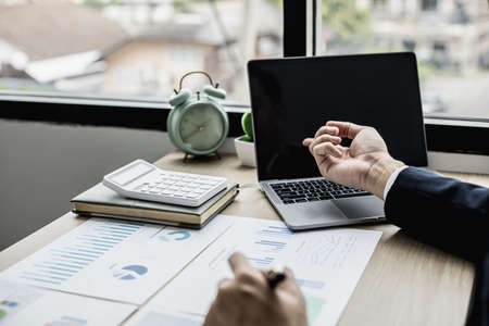 Man pointing at laptop screen and documents lying on the table, business man in black suit is sitting checking information from documents, he is a company executive. Company management concept.の写真素材