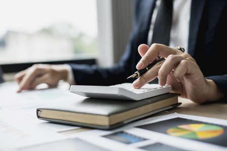 A man presses a white calculator to calculate numbers on a document, a businessman is sitting in his company's private office, he runs a startup company, he's a young man starting a business.の写真素材