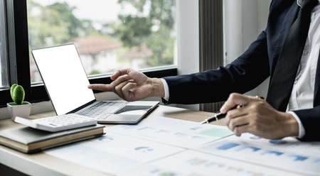Man pointing at laptop screen and documents lying on the table, business man in black suit is sitting checking information from documents, he is a company executive. Company management concept.の写真素材