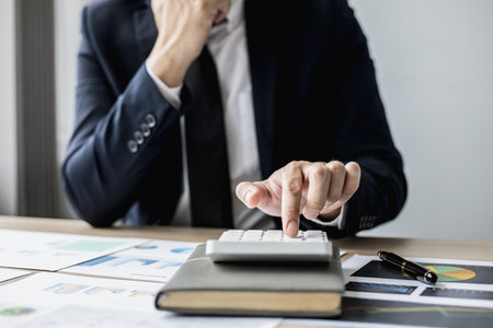 A man in a suit is pressing a calculator, a business man sitting in a private office at a company, he runs a startup company, he is a young man who starts a business. Startup company concept.の写真素材
