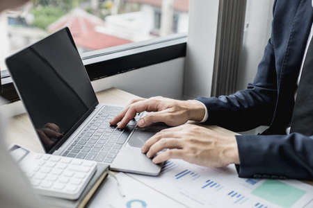 Man in suit sitting at work and using a laptop, he is a young businessman who founded a startup, he is typing text messages with his partner through an online messenger on his laptop.の写真素材