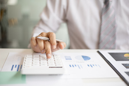 A business man is using a calculator to calculate company financial figures from earnings papers, a businessman sitting in his office where the company financial chart is placed. Financial concept.の写真素材