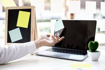 A businessman pointing at the screen of his laptop, on his desk there was a small board with small notepads, a laptop, a fake tree head in one tree.の写真素材