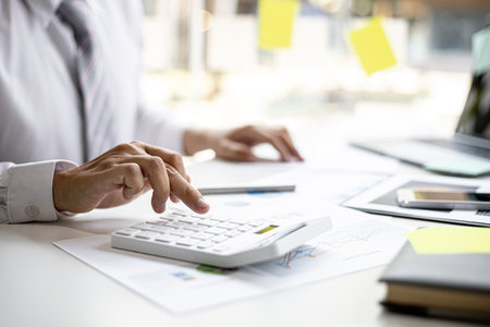 A business man is using a calculator to calculate company financial figures from earnings papers, a businessman sitting in his office where the company financial chart is placed. Financial concept.の写真素材
