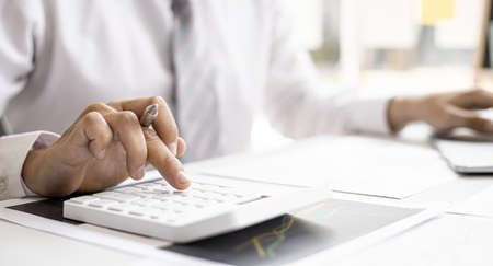A business man is using a calculator to calculate company financial figures from earnings papers, a businessman sitting in his office where the company financial chart is placed. Financial concept.の写真素材