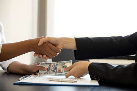 Two business women are shaking hands after negotiating a business jointly and giving away a large sum of dollars, a handshake is a Western greeting or congratulation. The concept of corruption.の写真素材