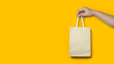 Close-up of a hand holding a shopping paper bag, using a paper bag instead of a plastic bag to help reduce global warming and plastic degradation. Isolated on a yellow backgroundの写真素材
