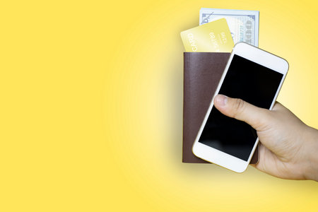 Close up Hand holding a brown passport and a mobile phone with a dollar bill and gold credit card inside, yellow background, passport used for international travelの写真素材