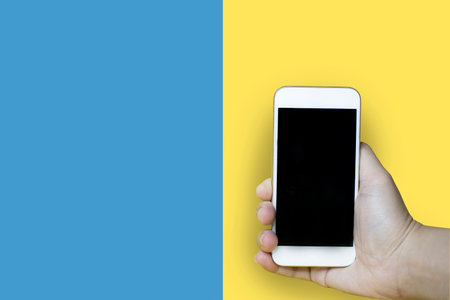 Close-up of a hand holding a mobile phone with a blue-yellow background, used for advertising photosの写真素材