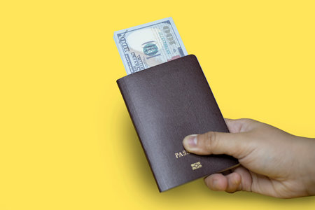 Close up Hand holding a brown passport with dollar bills inside, yellow background, passport used for international travel, Isolated on yellow backgroundの写真素材