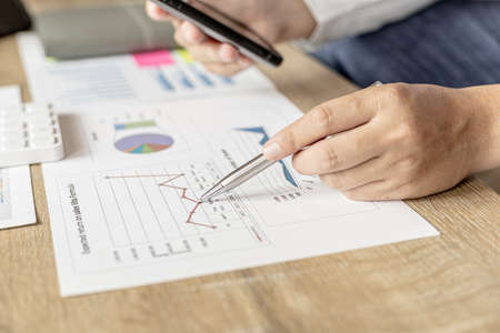 Businessman talking to a partner in a smartphone app, he is checking the accuracy of company financial data, financial status documents in line and pie charts. Finance concept.の写真素材