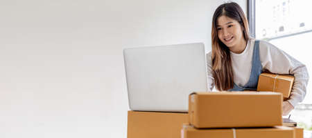 Owner of the online store carries two parcel boxes, she is checking the order balance from her laptop and preparing for delivery, a parcel for packing the goods. Online selling and online shopping concepts.の写真素材