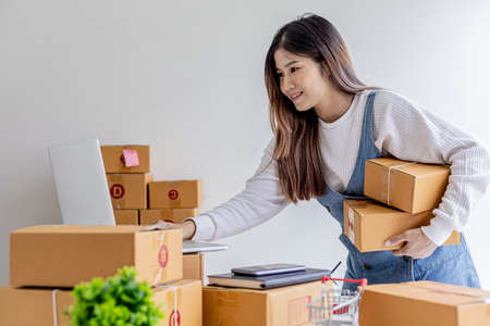 Owner of the online store carries two parcel boxes, she is checking the order balance from her laptop and preparing for delivery, a parcel for packing the goods. Online selling and online shopping concepts.の写真素材