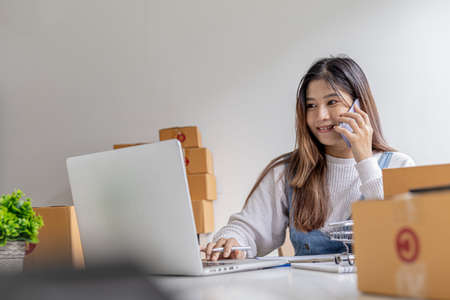 A woman talks on the phone and uses a laptop to confirm orders from customers on the website, packs the goods in parcels and sends them through a private courier company. Online shopping concept.の写真素材