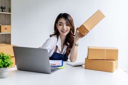 Asian woman holding parcel boxes and typing messages on laptop, she owns an online store, she is confirming orders from customers who order from online sites. Online selling concept.の写真素材