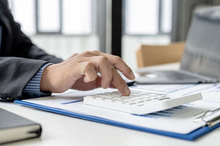 A businessman is pressing a calculator to check numbers on documents, he owns a company, he is checking company financial documents in his office, financial statements. Financial management concept.の写真素材