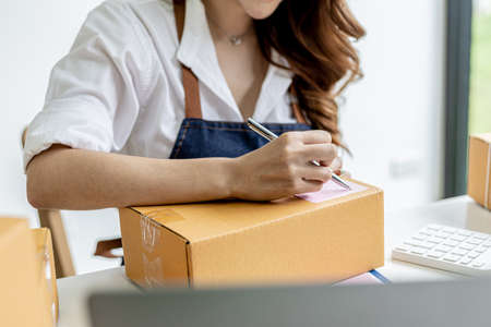 Asian woman writing customer's shipping information on parcel box, she owns an online store, she ships products to customers through a private courier company. Online selling concept.の写真素材