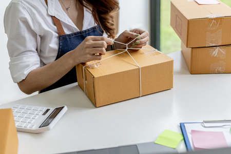 An Asian woman tying a parcel to a customer's box, she owns an online store, she packs and ships through a private transport company. Online selling and online shopping concepts.の写真素材