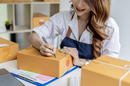 Asian woman writing customer's shipping information on parcel box, she owns an online store, she...