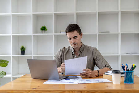 A young businessman sitting in the office working with a laptop, he is checking financial documents, he founded a startup, he is a young entrepreneur who has the skills to run a growing company.の写真素材