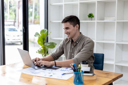 Young businessman sitting in the office with a laptop, typing messages with partners through chat, he founded a startup, he is a young businessman who is skilled in running a growing company.の写真素材