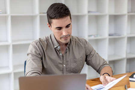 A young businessman sitting in the office working with a laptop, he is checking financial documents, he founded a startup, he is a young entrepreneur who has the skills to run a growing company.の写真素材