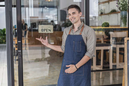 A male cafe employee standing in front of the store greets customers and has a sign that says open to show that the shop is open, a male employee opens the shop to serve food and beverages.の写真素材