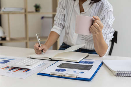 Businesswoman taking notes in a notebook and drinking coffee, she is checking product sales and financial documents, she is the executive of a startup company. Business administration concept.の写真素材