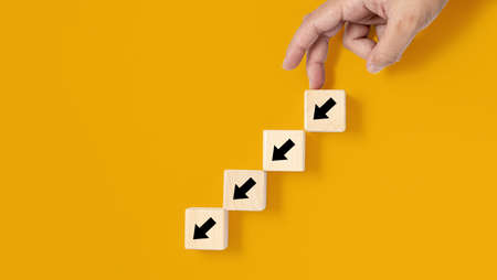 A square wooden block is placed on a yellow background, on the wooden block showing a down arrow and a hand doing a downward movement. banner with copy space for text, poster, mockup template.の写真素材