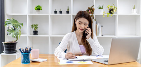 Beautiful Asian businesswoman sitting in her room, talking to her partner via mobile phone and checking financial documents, she is a female executive of a startup company. Financial management.の写真素材