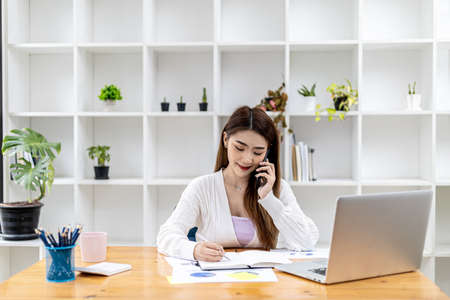 Beautiful Asian businesswoman sitting in her room, talking to her partner via mobile phone and checking financial documents, she is a female executive of a startup company. Financial management.の写真素材