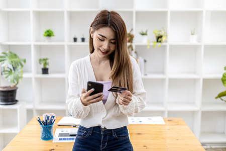 Beautiful Asian woman standing with smart phone and credit card, she fills in credit card information to pay for goods and services, online shopping concept pay by credit card and use via smartphone.の写真素材