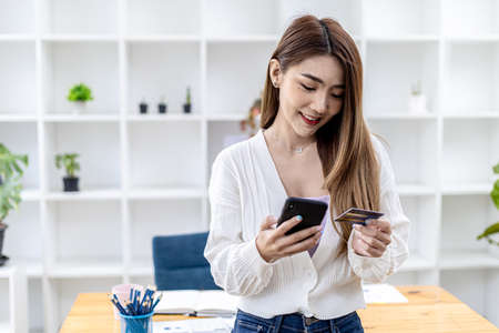 Beautiful Asian woman standing with smart phone and credit card, she fills in credit card information to pay for goods and services, online shopping concept pay by credit card and use via smartphone.の写真素材