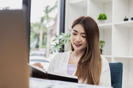 A beautiful Asian businesswoman working in the room, she is writing her schedule to remind herself in her notebook, she is a female executive of a startup company. Concept of financial management.の写真素材