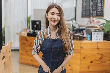 Portrait of a beautiful Asian woman in an apron standing in a coffee shop, she owns a coffee shop, the concept of a food and beverage business. Store management by a business woman.の写真素材