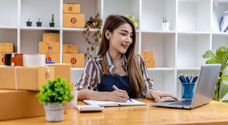 A beautiful Asian business owner opens an online store, she is checking orders from customers via laptop, sending goods through a courier company, concept of a woman opening an online business.の写真素材