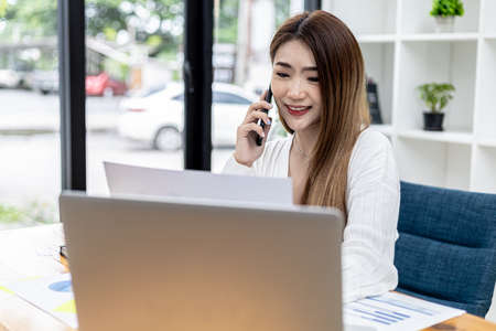 Beautiful Asian businesswoman sitting in her room, talking to her partner via mobile phone and checking financial documents, she is a female executive of a startup company. Financial management.の写真素材