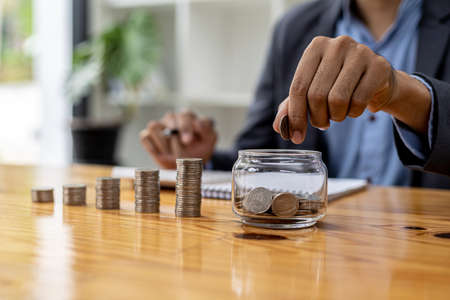 A man is dropping coins into a glass jar with a number of coins inside, on a desk are stacked coins from low to high showing the growth of money. Concept of saving and investing money.の写真素材