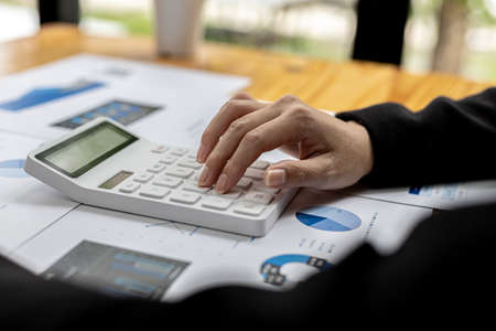 Close-up of a businesswoman using a white calculator, a financial businessman examining the numerical data on a company financial document, she uses a calculator to verify the accuracy of numbers.の写真素材