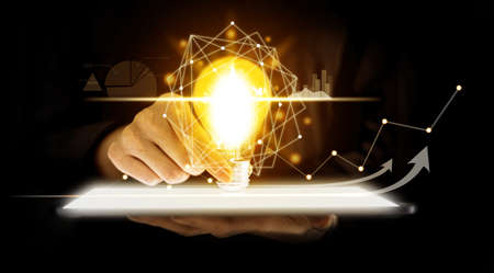 A person holding a light bulb placed on a tablet screen, graphic charts and fluctuations in...