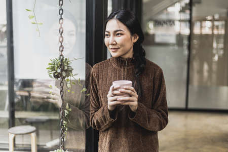 A beautiful Asian woman showing a smiling face, she holds a cup of coffee and looks out the window of her office to relax after a long time at work. Concept of relaxing after work.の写真素材
