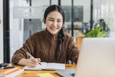 A beautiful Asian woman shows a smiling face and sitting at her desk working, she is checking company financial documents, she is a female executive of a startup company.の写真素材