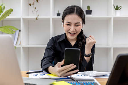 Woman holding a smartphone and gesturing, Asian business woman showing joy after watching the company's growing earnings in the past month, she manages the growth. Business administration concept.の写真素材